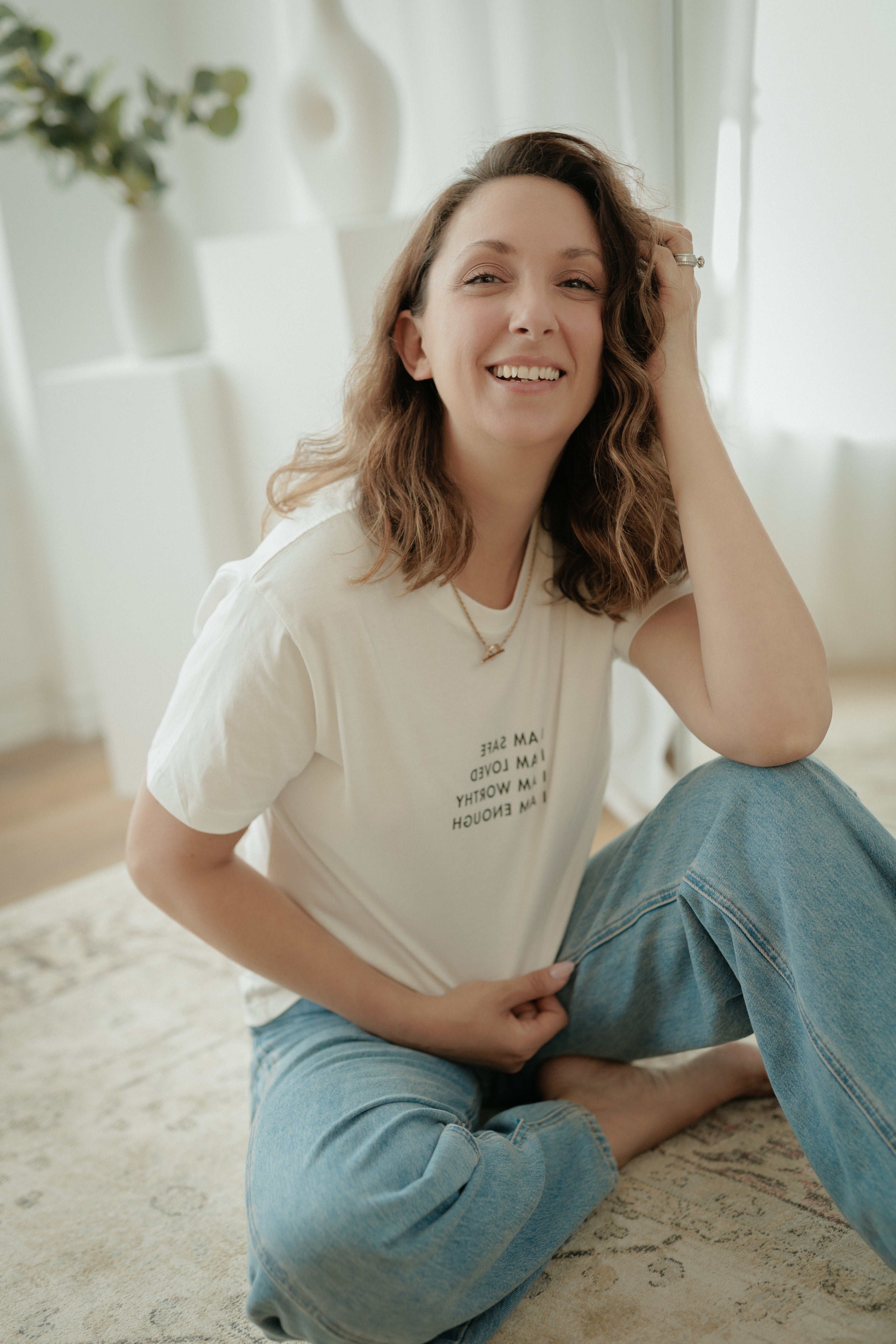 A woman sitting on a patterned rug in a bright, modern room, wearing a cream-colored graphic t-shirt and light blue jeans.