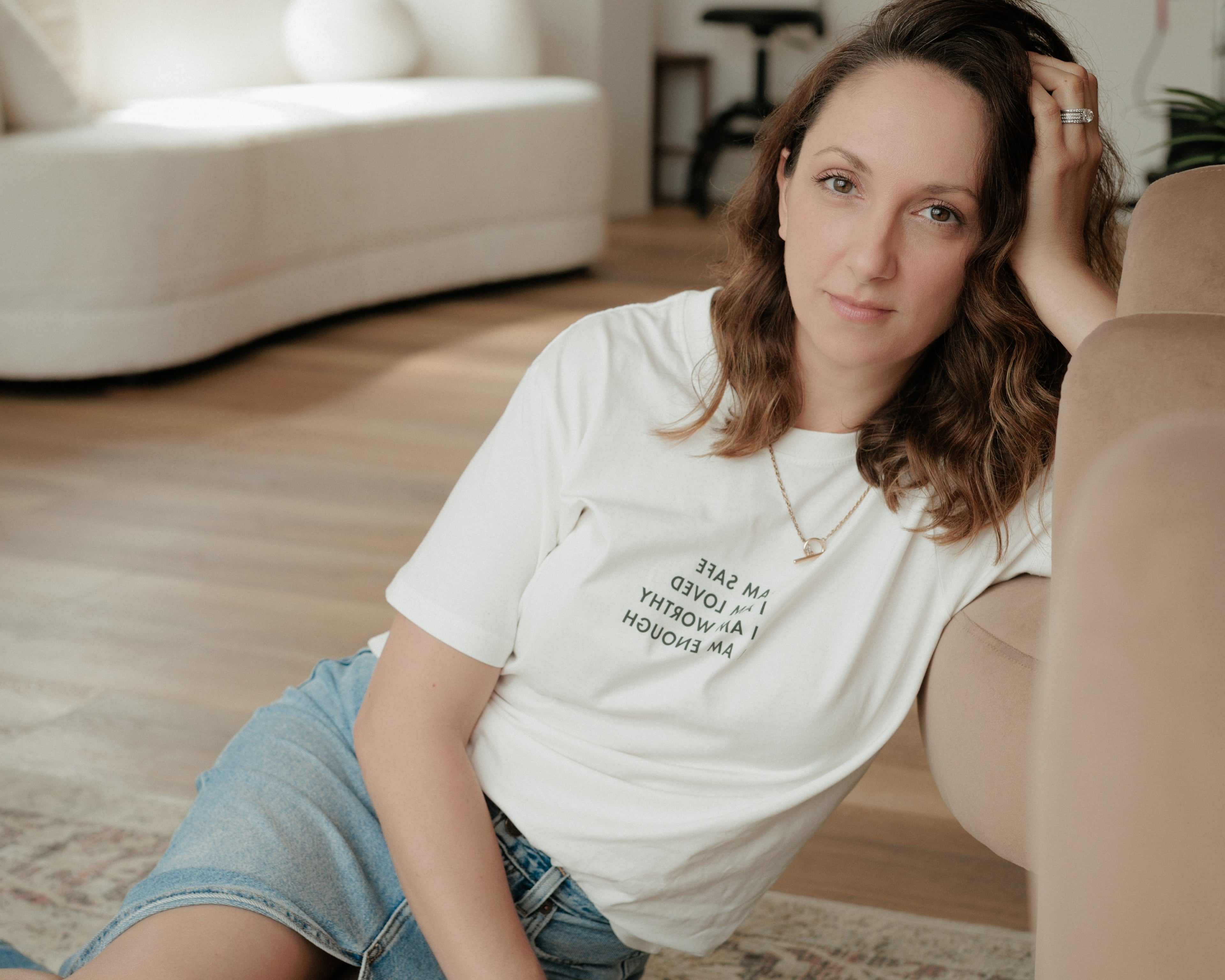 A woman wearing a white graphic t-shirt and denim skirt sits on a patterned rug in a modern living room setting.