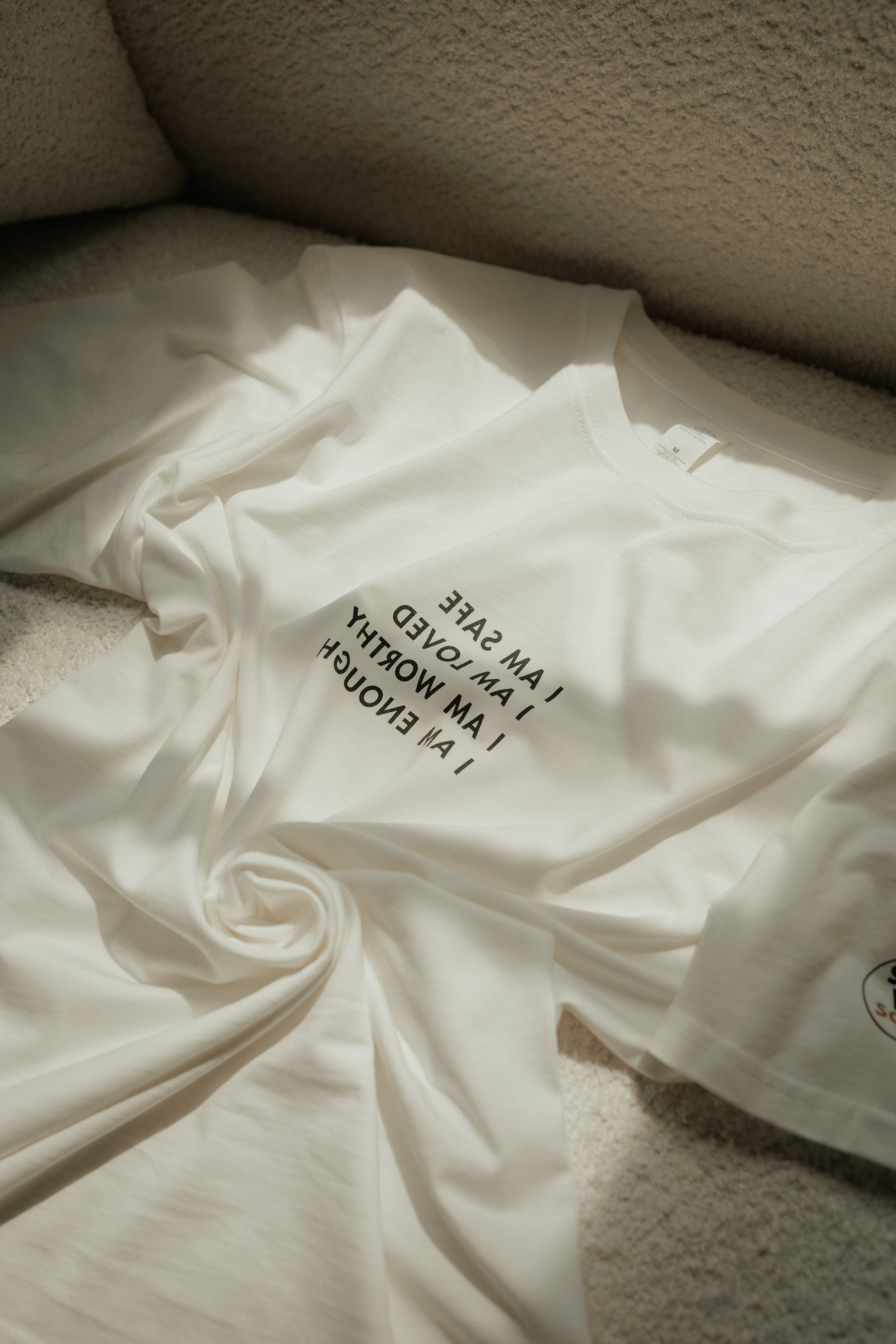 A white short-sleeve T-shirt with black text reading 'I AM WORTHY I AM SAFE I AM LOVED I AM ENOUGH' arranged in a reversed design on the chest, draped on a textured beige surface.